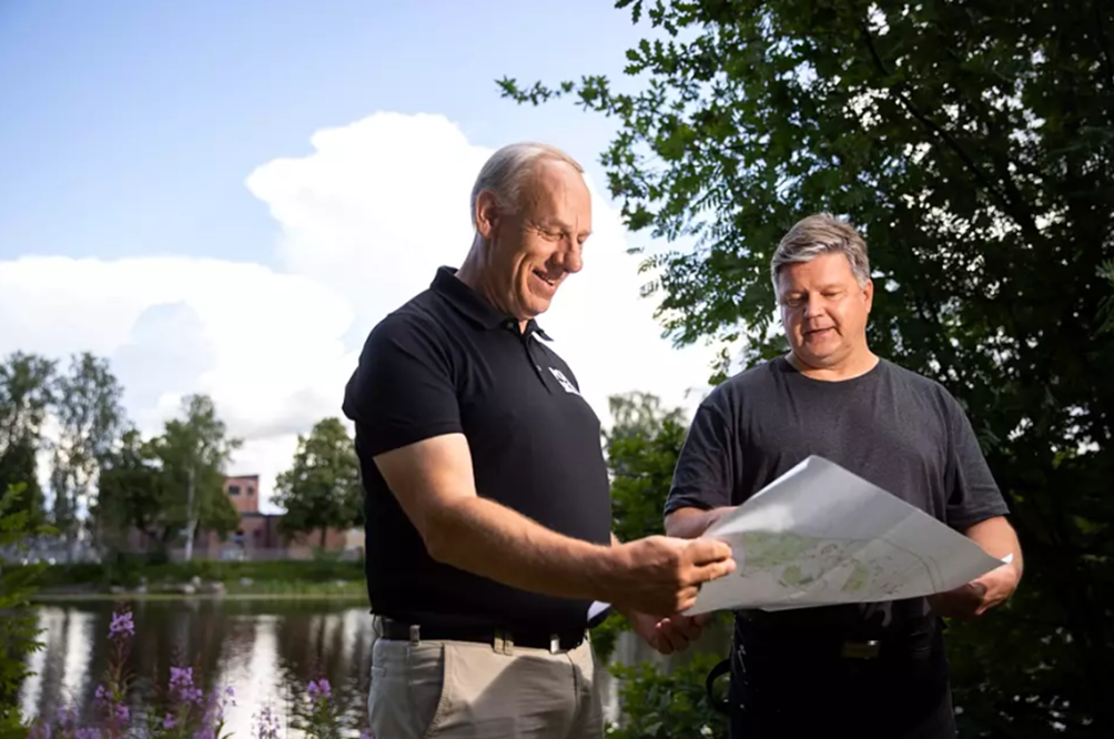 Kari Sarpela ja Tero Isoviita tarkistavat kytkentäkaaviot. Taustalla näkyvä jokimaiseman maamerkki, Isosannan sähköasema