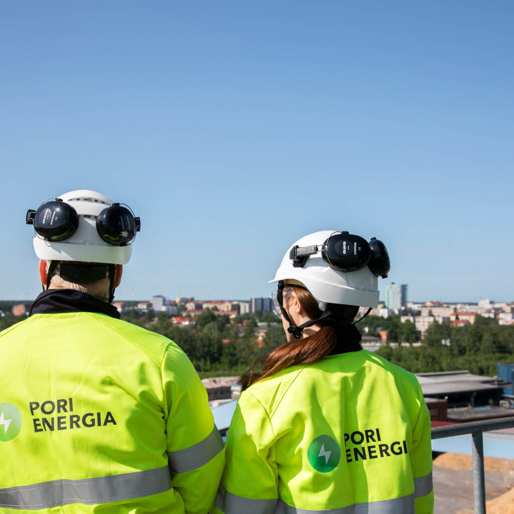Kaksi Pori Energian työvarusteisiin pukeutunutta henkilöä seisoo korkealla tasanteella Aittaluodon voimalaitoksen ulkoympäristössä selkä kameraan päin