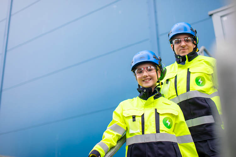 Kaksi Pori Energian työvarusteisiin pukeutunutta henkilöä seisoo rappusissa Aittaluodon voimalaitoksen ulkoympäristössä ja katsoo kameraan
